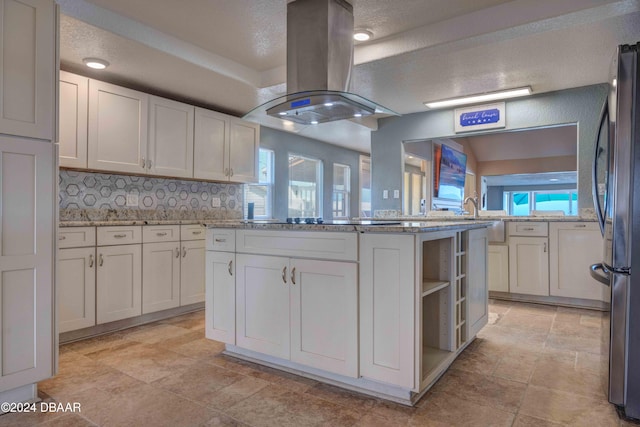 kitchen with white cabinetry, island exhaust hood, light stone counters, black fridge, and decorative backsplash