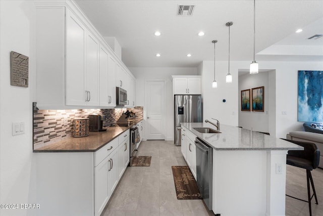 kitchen featuring white cabinetry, appliances with stainless steel finishes, dark stone counters, hanging light fixtures, and a kitchen island with sink