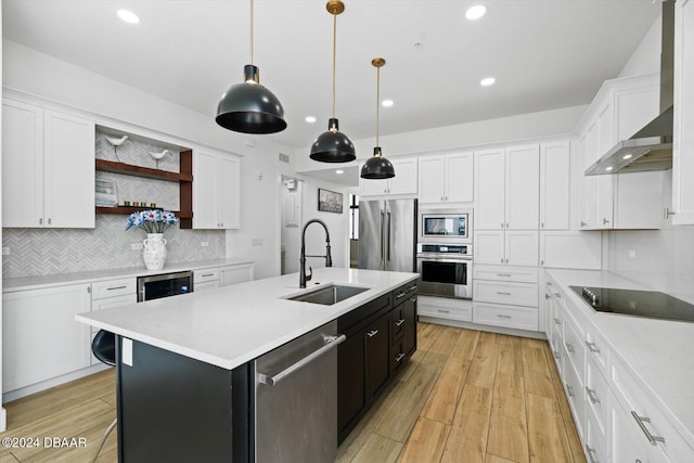 kitchen featuring a center island with sink, stainless steel appliances, decorative light fixtures, wall chimney exhaust hood, and white cabinets