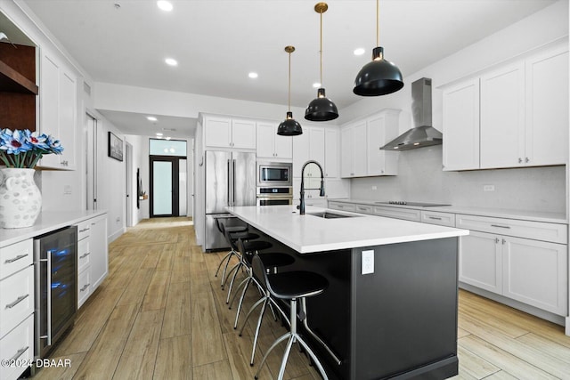 kitchen with appliances with stainless steel finishes, wall chimney range hood, wine cooler, sink, and a center island with sink