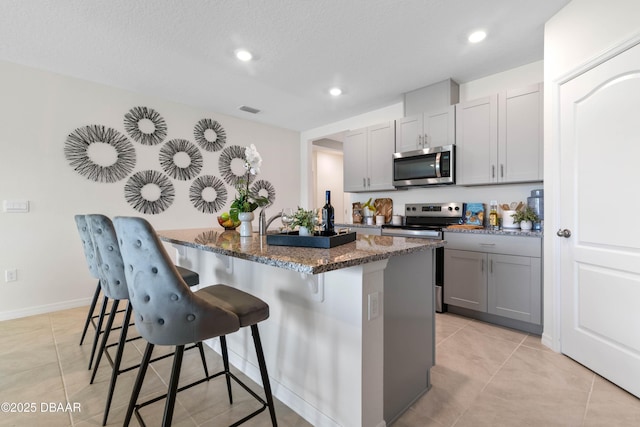 kitchen featuring a kitchen bar, gray cabinetry, dark stone counters, stainless steel appliances, and a kitchen island with sink