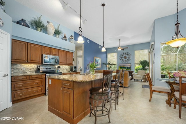 kitchen with pendant lighting, decorative backsplash, light stone counters, stainless steel appliances, and a center island with sink