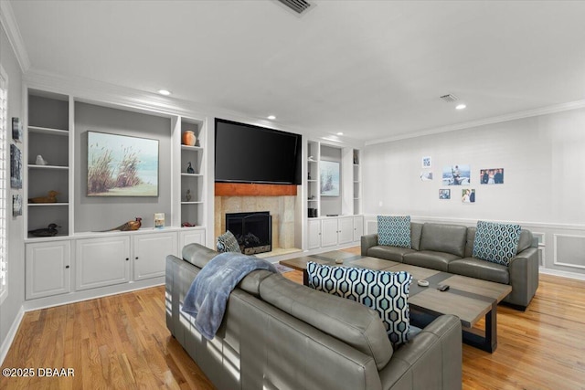 living room with built in shelves, light wood-type flooring, a tile fireplace, and crown molding