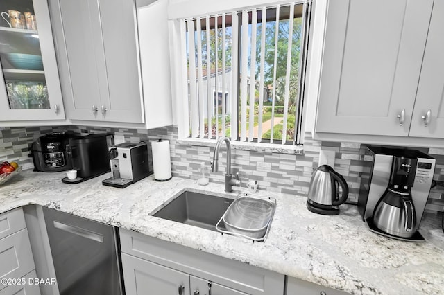 kitchen featuring backsplash, light stone countertops, dishwasher, a sink, and glass insert cabinets