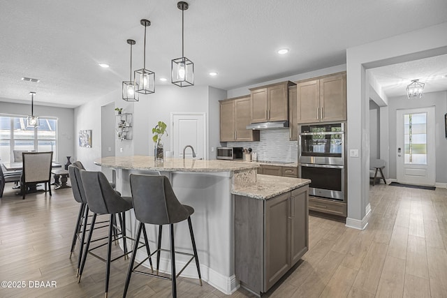 kitchen featuring a large island, backsplash, appliances with stainless steel finishes, light wood-type flooring, and under cabinet range hood