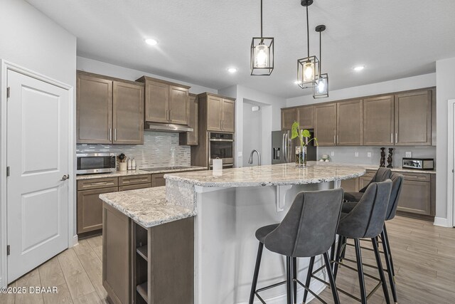 kitchen featuring light wood-style floors, a breakfast bar area, stainless steel appliances, under cabinet range hood, and backsplash