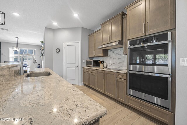 kitchen with under cabinet range hood, a sink, appliances with stainless steel finishes, light wood-type flooring, and backsplash