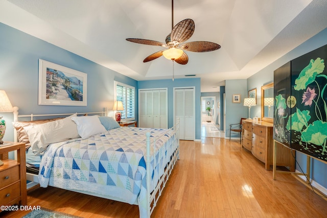 bedroom featuring a raised ceiling, multiple closets, ceiling fan, and light hardwood / wood-style flooring