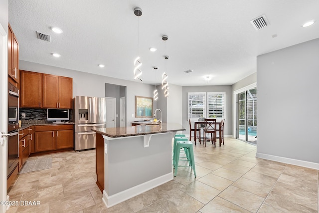 kitchen featuring stainless steel appliances, visible vents, decorative backsplash, dark stone counters, and brown cabinetry