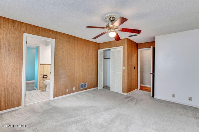 unfurnished bedroom with light colored carpet, a textured ceiling, wooden walls, ensuite bathroom, and ceiling fan