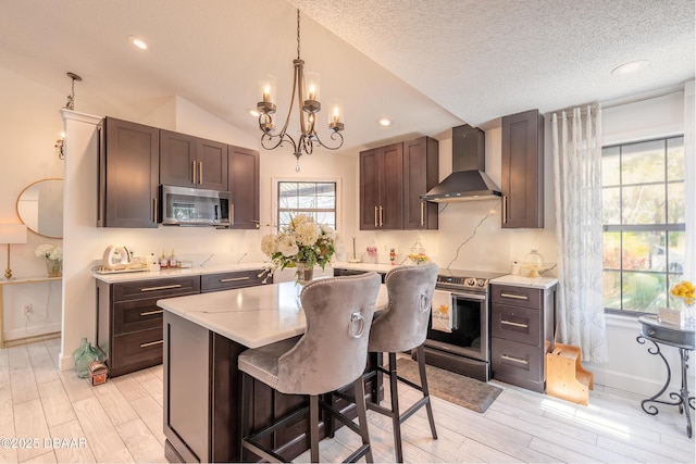 kitchen featuring wall chimney range hood, an inviting chandelier, appliances with stainless steel finishes, light wood finished floors, and light countertops