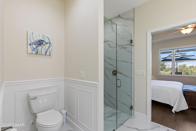 ensuite bathroom featuring ceiling fan, toilet, a decorative wall, wainscoting, and a marble finish shower