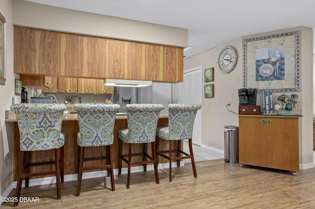 kitchen with kitchen peninsula, stainless steel refrigerator with ice dispenser, light wood-type flooring, a breakfast bar, and exhaust hood