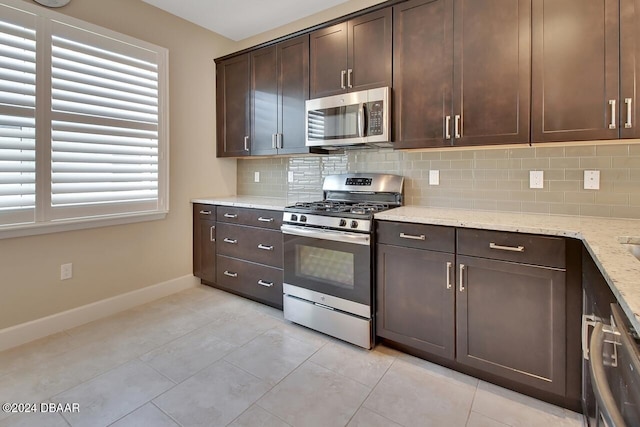 kitchen featuring dark brown cabinetry, light stone counters, backsplash, light tile patterned flooring, and appliances with stainless steel finishes