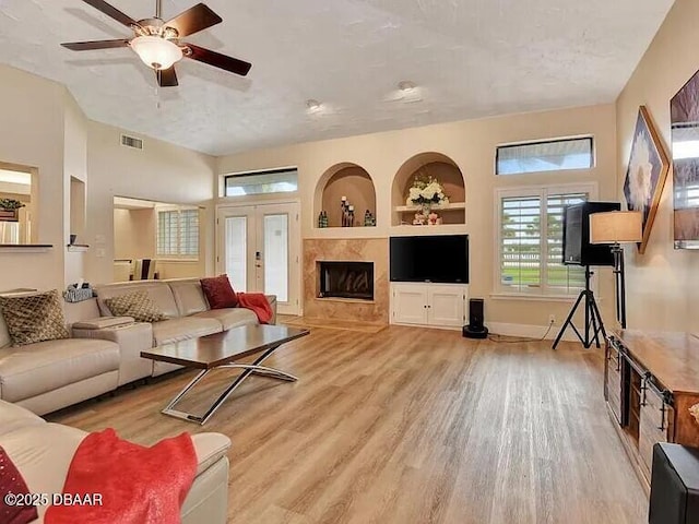 living room featuring built in features, light hardwood / wood-style flooring, a tiled fireplace, ceiling fan, and french doors