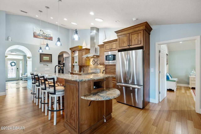 kitchen with wall chimney exhaust hood, light hardwood / wood-style flooring, a large island, appliances with stainless steel finishes, and decorative light fixtures