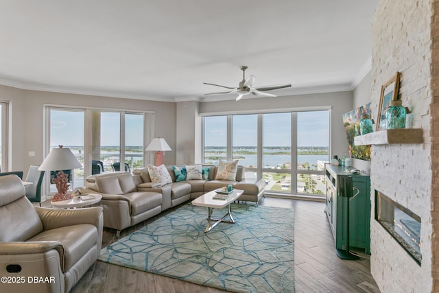 living room with ceiling fan, a stone fireplace, hardwood / wood-style floors, a water view, and ornamental molding