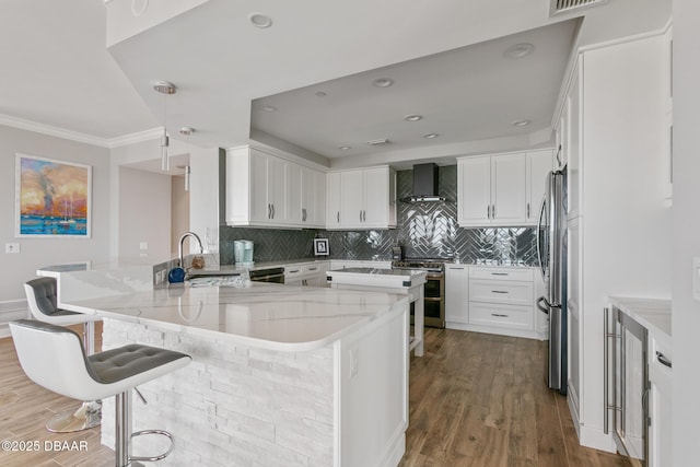 kitchen featuring a kitchen breakfast bar, stainless steel appliances, wall chimney range hood, sink, and white cabinetry