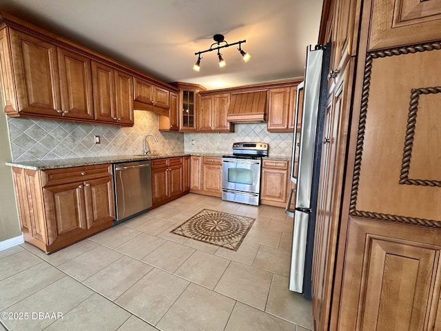 kitchen with brown cabinetry, light stone countertops, stainless steel appliances, premium range hood, and a sink