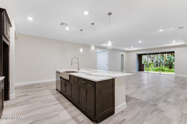 kitchen with sink, hanging light fixtures, a center island with sink, a raised ceiling, and light stone countertops