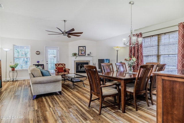 dining area featuring a tiled fireplace, hardwood / wood-style floors, ceiling fan with notable chandelier, and vaulted ceiling