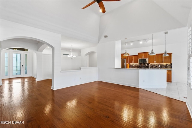 unfurnished living room with sink, ceiling fan with notable chandelier, high vaulted ceiling, and light hardwood / wood-style floors