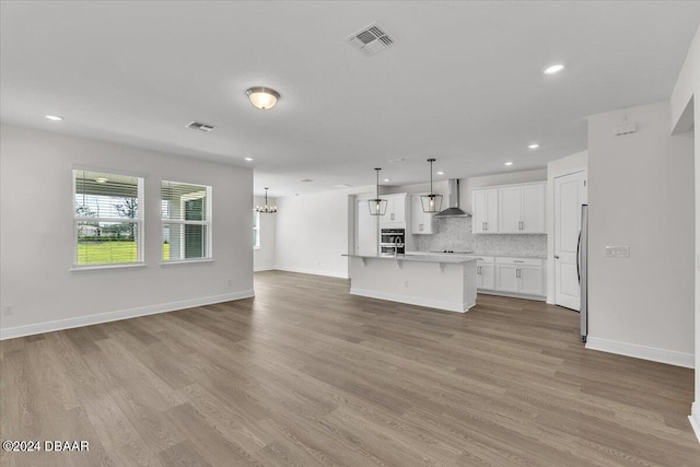 kitchen with white cabinetry, hanging light fixtures, wall chimney range hood, light hardwood / wood-style flooring, and a kitchen island with sink