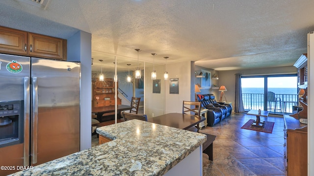 kitchen with stainless steel refrigerator with ice dispenser, light stone counters, a textured ceiling, a water view, and decorative light fixtures