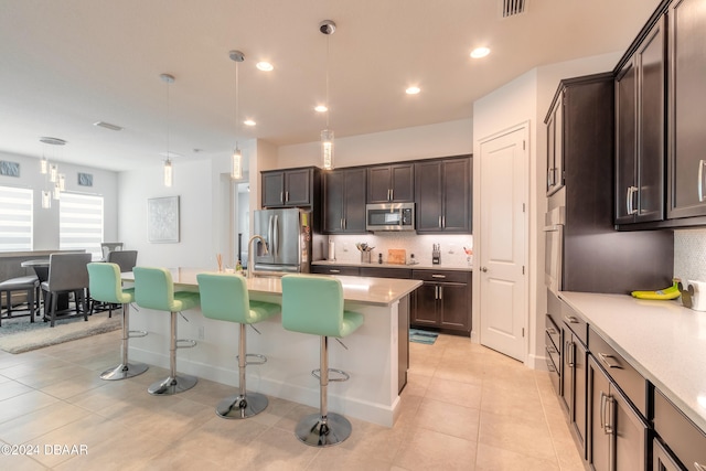 kitchen featuring stainless steel appliances, dark brown cabinetry, hanging light fixtures, an island with sink, and a breakfast bar