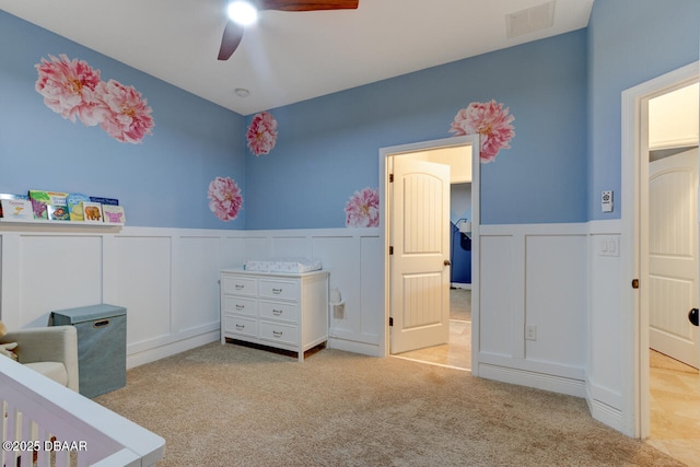 carpeted bedroom featuring visible vents, a decorative wall, a ceiling fan, and wainscoting