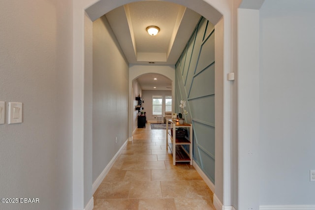 hallway with arched walkways, a raised ceiling, and baseboards