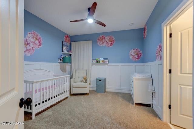 carpeted bedroom featuring a nursery area, a wainscoted wall, ceiling fan, and a decorative wall