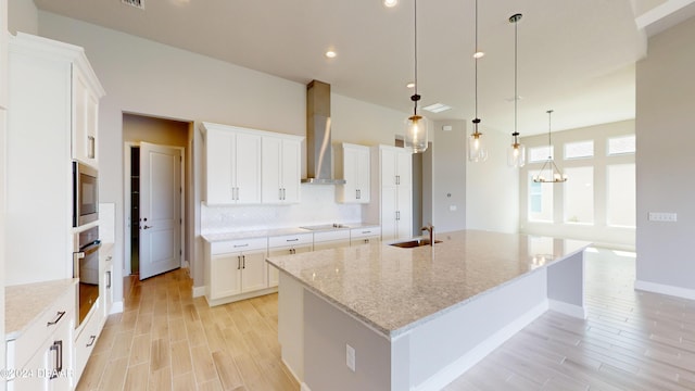 kitchen featuring a spacious island, wall chimney range hood, light stone countertops, white cabinetry, and decorative light fixtures