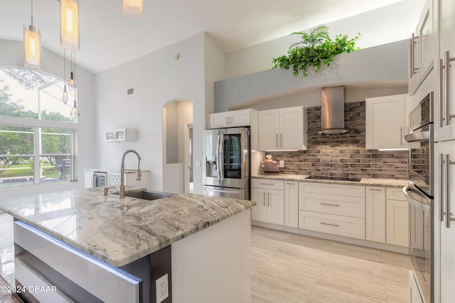 kitchen with light stone counters, wall chimney range hood, stainless steel fridge with ice dispenser, pendant lighting, and a kitchen island with sink