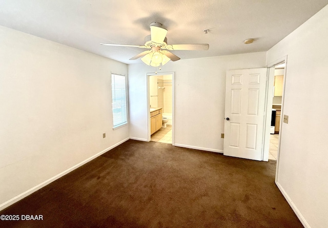 unfurnished bedroom featuring ensuite bath, baseboards, dark carpet, and a ceiling fan