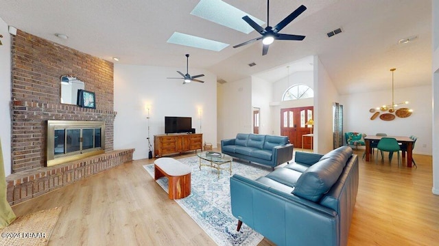 living room with light hardwood / wood-style flooring, high vaulted ceiling, a brick fireplace, a skylight, and ceiling fan with notable chandelier