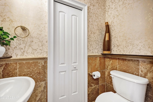 bathroom featuring toilet, tile walls, a closet, wainscoting, and wallpapered walls