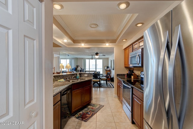 kitchen with appliances with stainless steel finishes, a raised ceiling, ceiling fan, and dark stone countertops