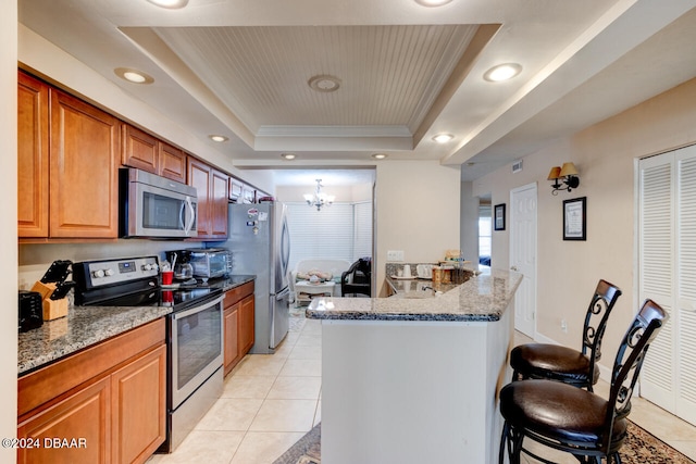 kitchen featuring appliances with stainless steel finishes, a kitchen breakfast bar, dark stone counters, a raised ceiling, and a notable chandelier