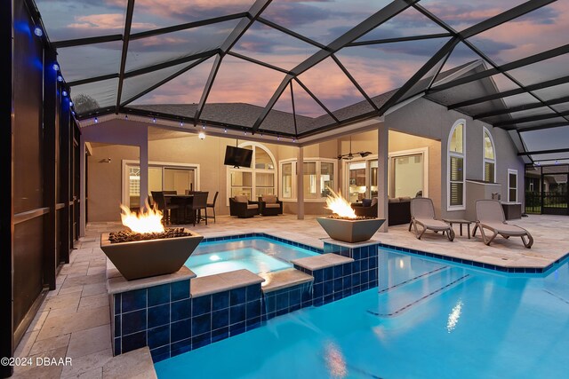 pool at dusk with an in ground hot tub, an outdoor living space with a fire pit, ceiling fan, glass enclosure, and a patio area