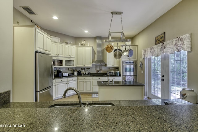 kitchen with sink, wall chimney exhaust hood, hanging light fixtures, stainless steel appliances, and dark stone countertops