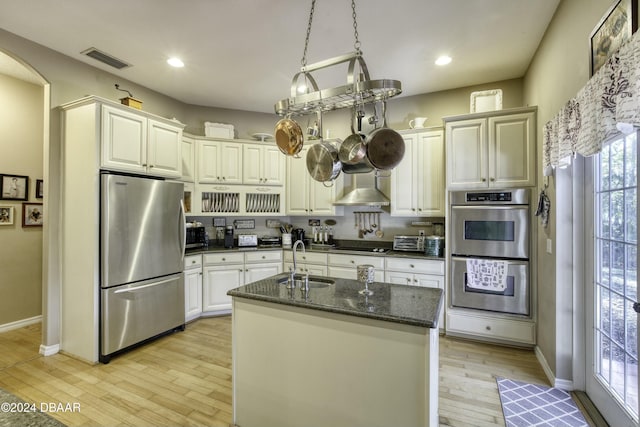 kitchen with wall chimney exhaust hood, stainless steel appliances, sink, a center island with sink, and dark stone countertops