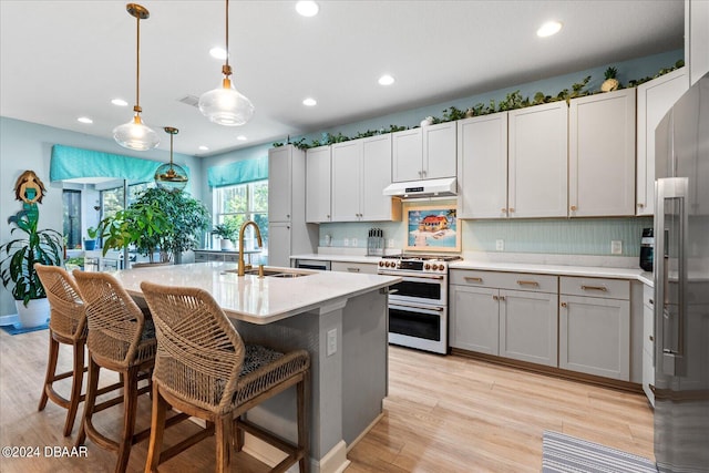 kitchen with stainless steel appliances, light wood-type flooring, decorative light fixtures, sink, and an island with sink