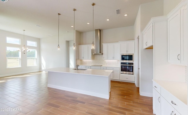 kitchen with wall chimney exhaust hood, stainless steel appliances, pendant lighting, a center island with sink, and white cabinetry