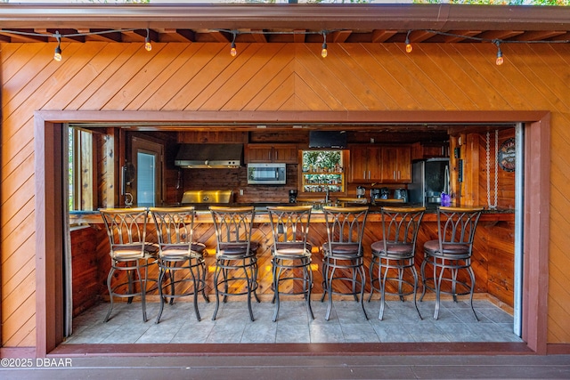 bar featuring stainless steel microwave, ventilation hood, wood walls, freestanding refrigerator, and range