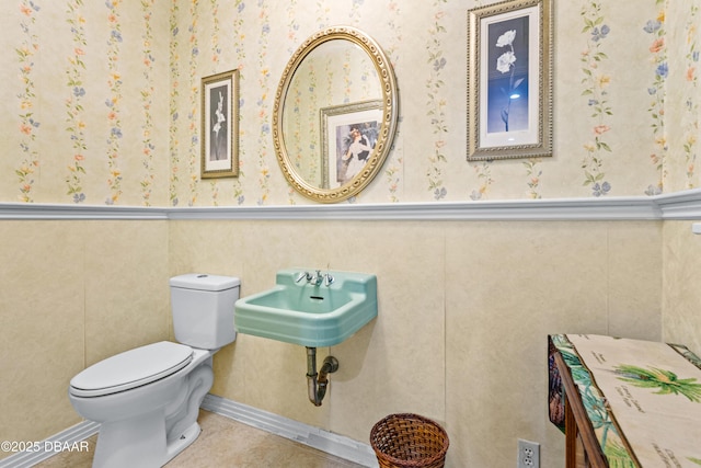 half bathroom with a wainscoted wall, wallpapered walls, a sink, tile patterned floors, and toilet