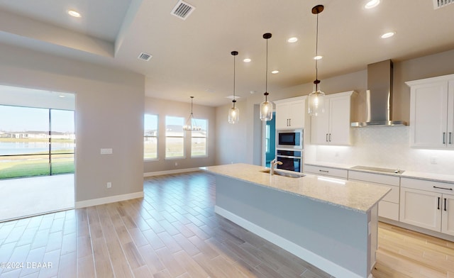 kitchen featuring wall chimney range hood, an island with sink, decorative light fixtures, white cabinetry, and stainless steel appliances