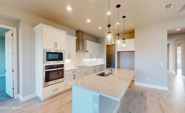 kitchen featuring wall chimney exhaust hood, decorative light fixtures, a center island with sink, white cabinets, and appliances with stainless steel finishes