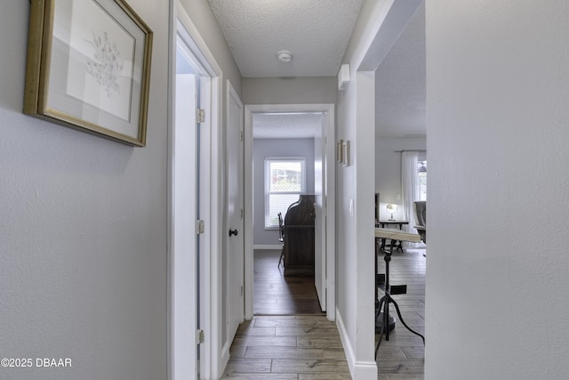 corridor featuring baseboards, wood finished floors, a textured wall, and a textured ceiling