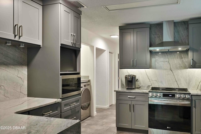 kitchen featuring washer / dryer, gray cabinetry, light stone counters, stainless steel appliances, and wall chimney range hood